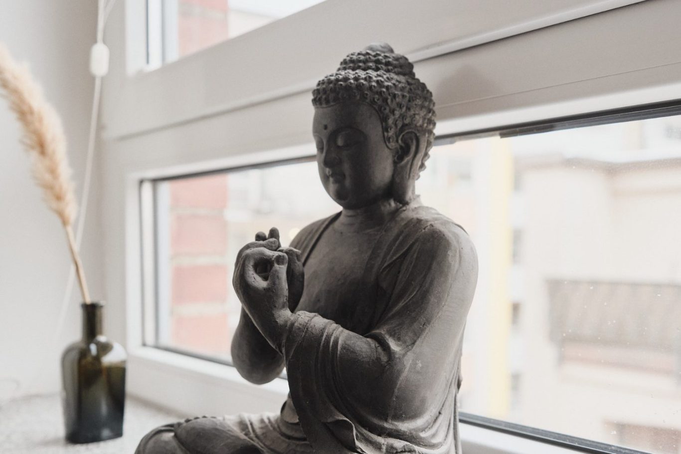 Buddha-Skulptur auf einem Fensterbrett, umgeben von Pflanzen und einer Flasche.
