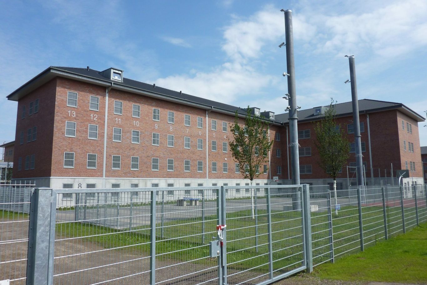 Ein dreigeschossiges, rotes Backsteingebäude umgeben von einem Zaun. Blaues Himmellicht.