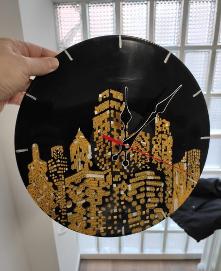 Wanduhr mit goldenem Stadtmotiv und roten Zeigern auf schwarzem Hintergrund.