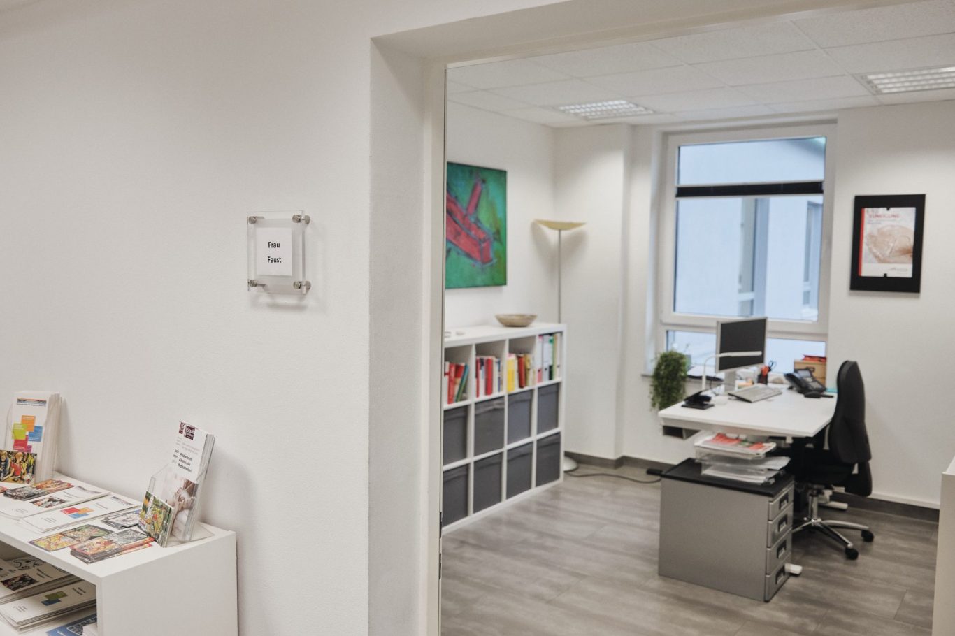 Heller Büroraum mit Schreibtisch, Computer und Regalen, dekoriert mit Bildern.